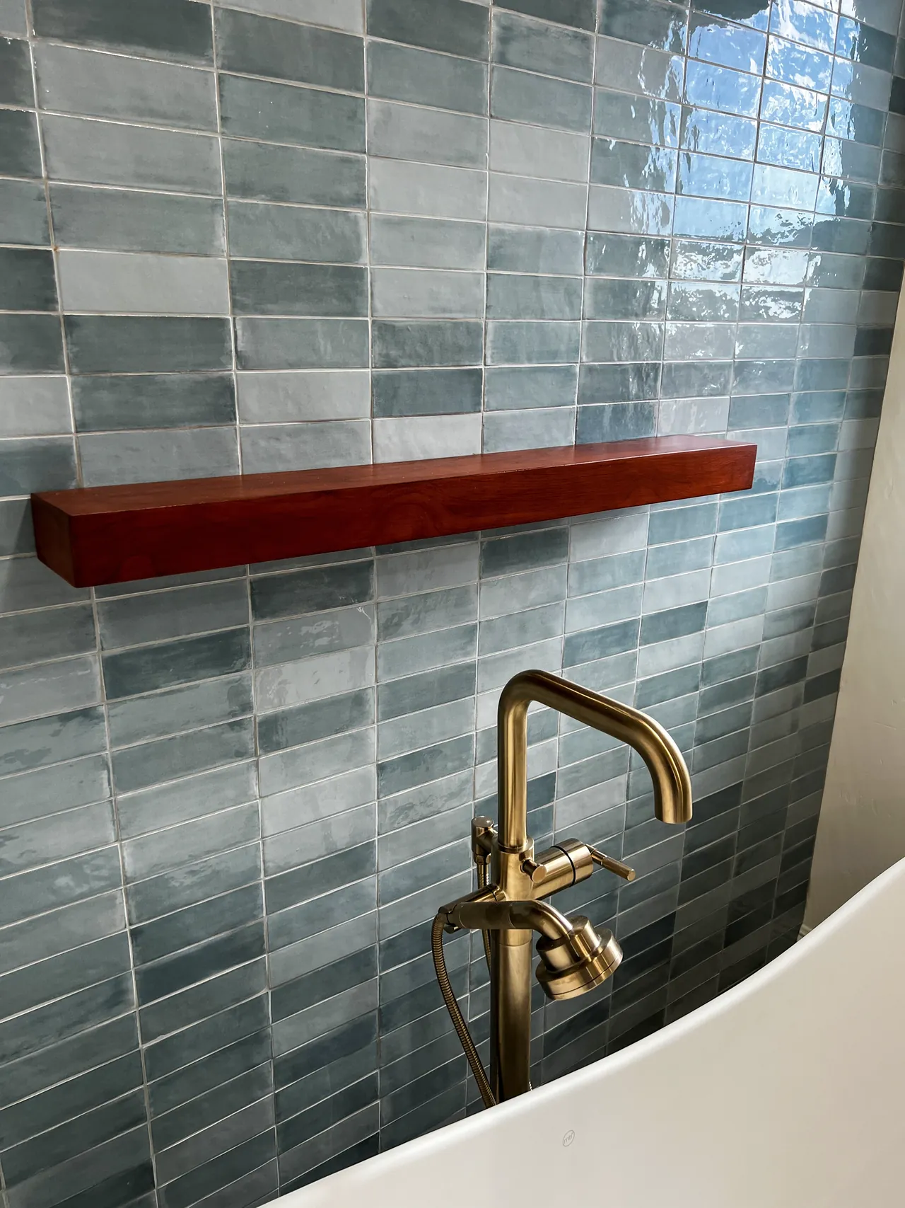 Brushed brass floor-mounted tub filler below a floating wood shelf on the blue tile wall