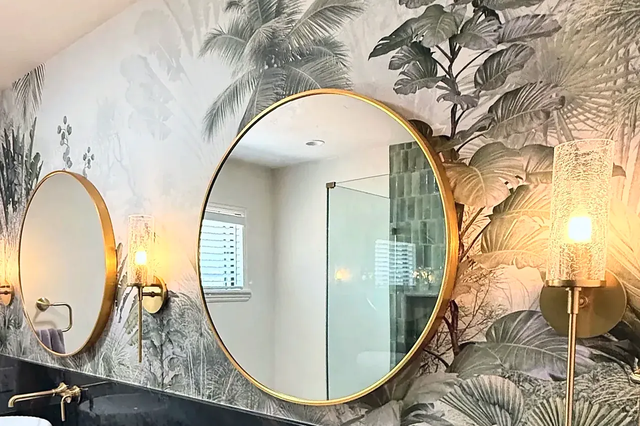 The finished vanity wall, with botanical wallpaper, two round brass mirrors and glass sconces