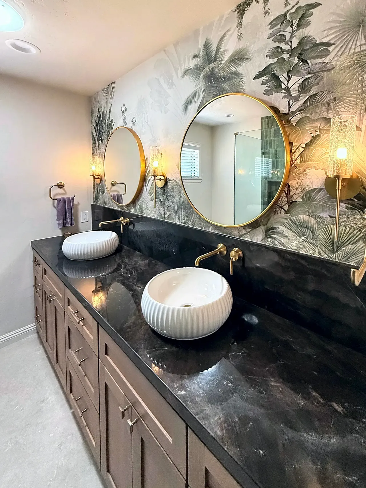 The finished vanity wall: round brass mirrors, vessel sinks, a dark stone top and botanical wallpaper