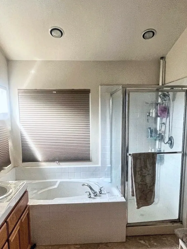 Primary bathroom before the remodel, with a drop-in tub in a white tile deck under the window and a framed glass shower stall
