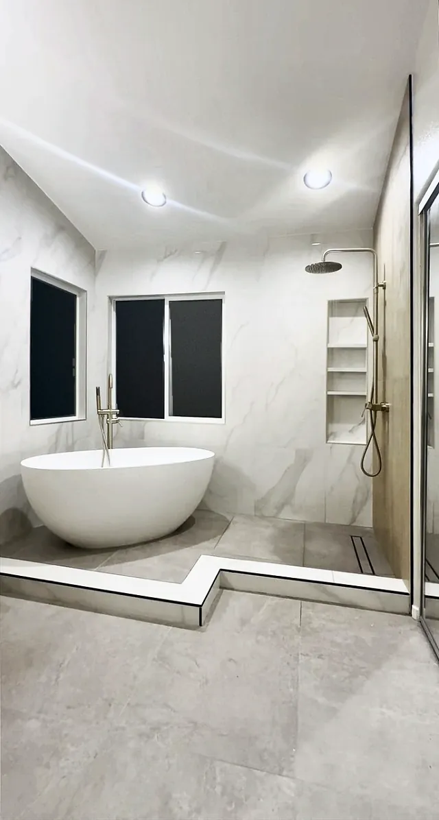 Freestanding soaking tub under two windows, marble-look tile to the ceiling, and a shower niche and rain head on the right