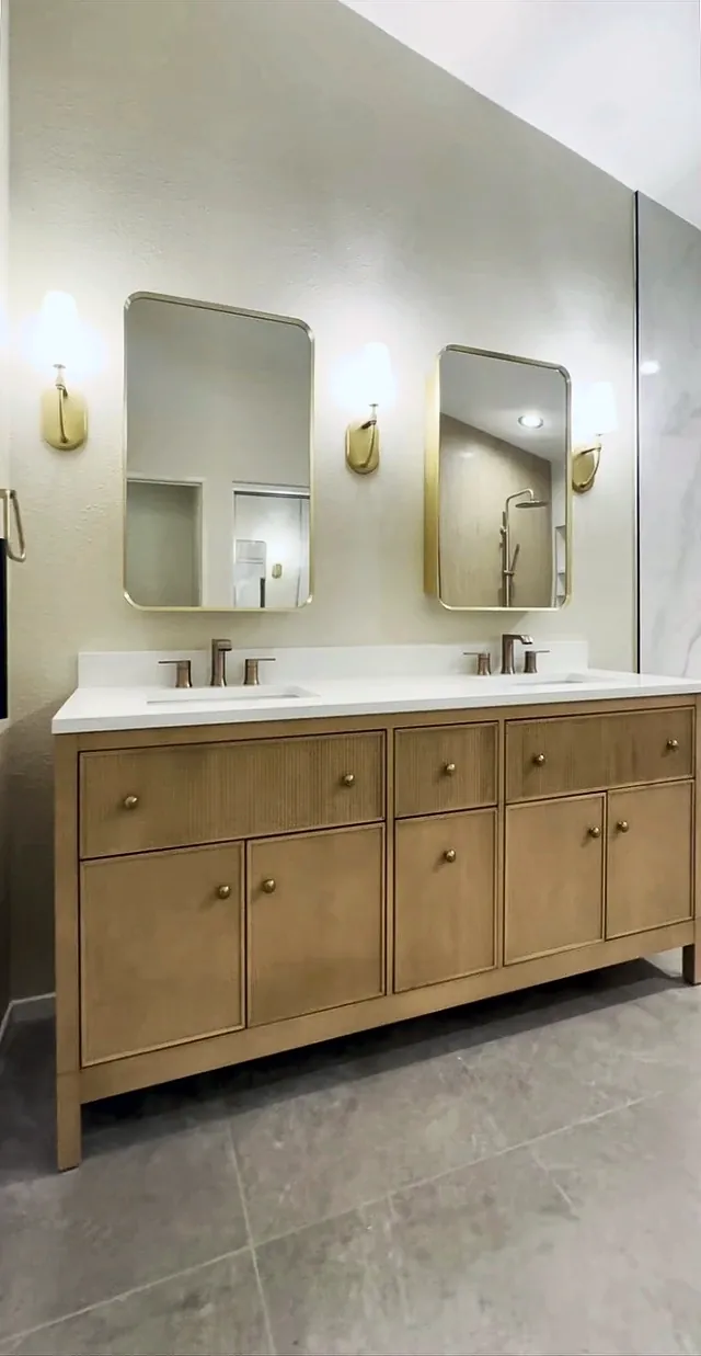Light oak double vanity with fluted drawer fronts, a white top, two brass framed mirrors and three sconces