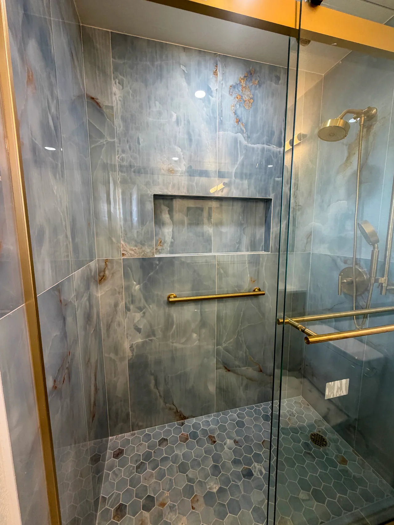 Walk-in shower in blue onyx-look porcelain with a wide niche, a brushed gold grab bar and a sliding glass door