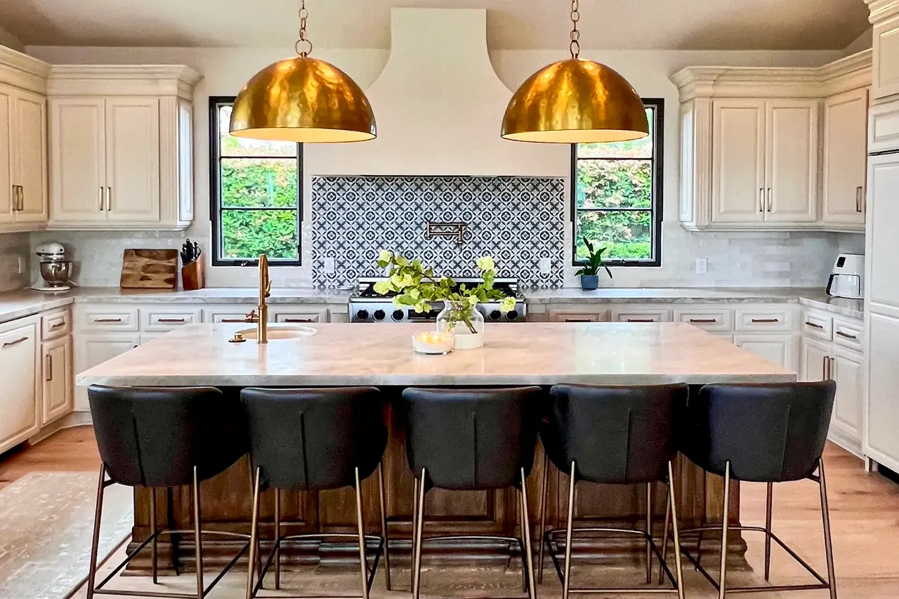 Finished kitchen with two hammered brass dome pendants over a large island, white raised-panel cabinets and a patterned tile backsplash between black-framed windows