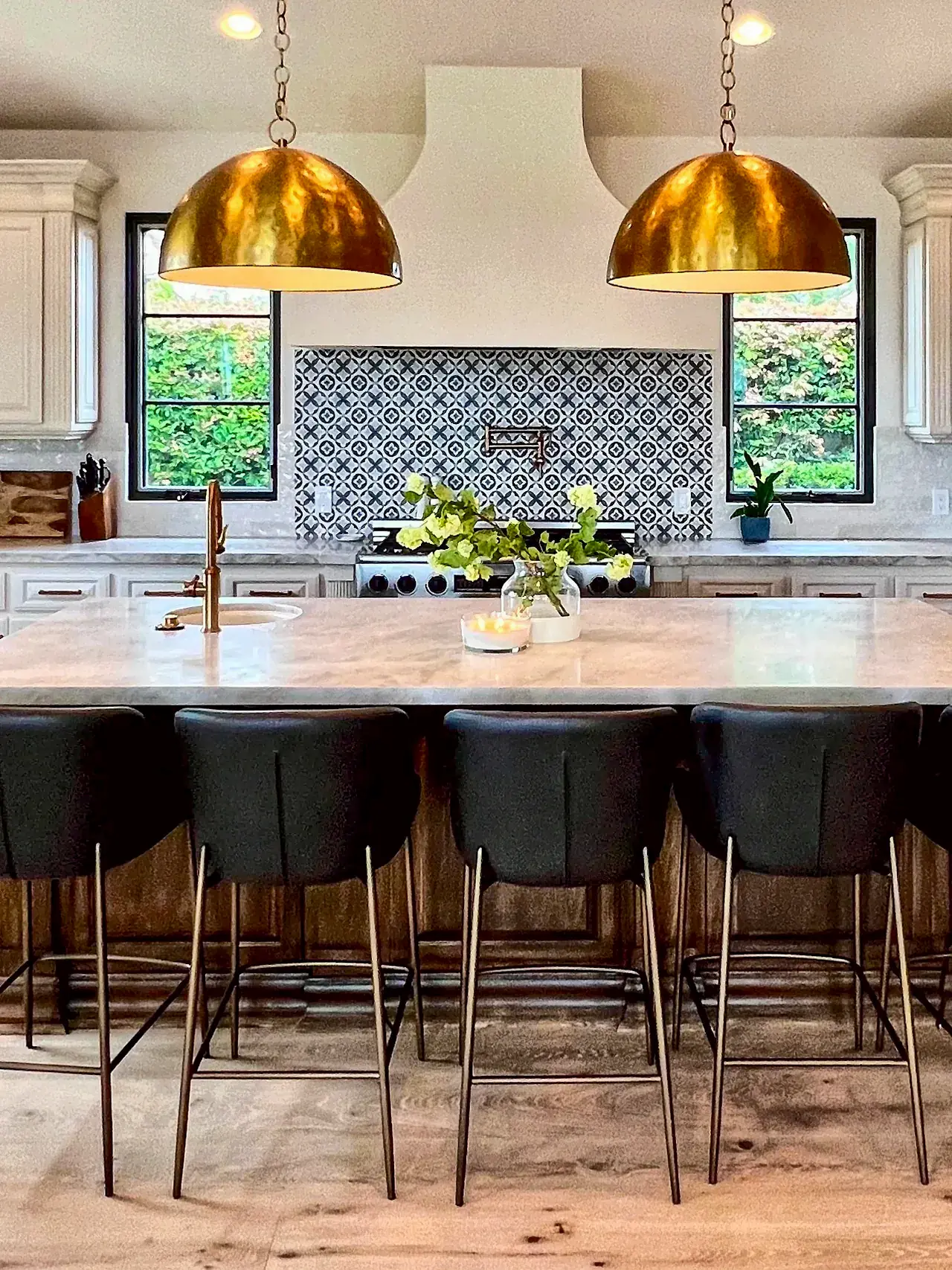 The same Santaluz kitchen after the remodel, with cream cabinets, a smooth white range hood, a patterned tile backsplash, black window frames, brass pendants, light wood floors and a new stone island top