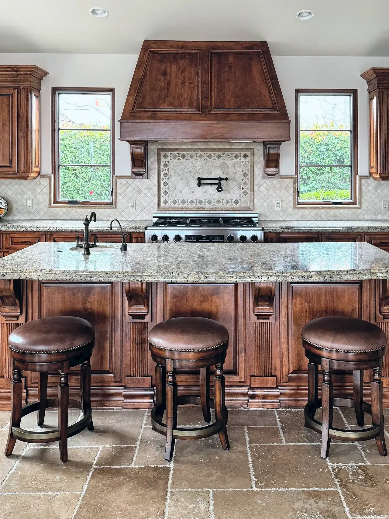 Kitchen before, with dark stained cabinets, a wood range hood, a granite island and tumbled stone floors