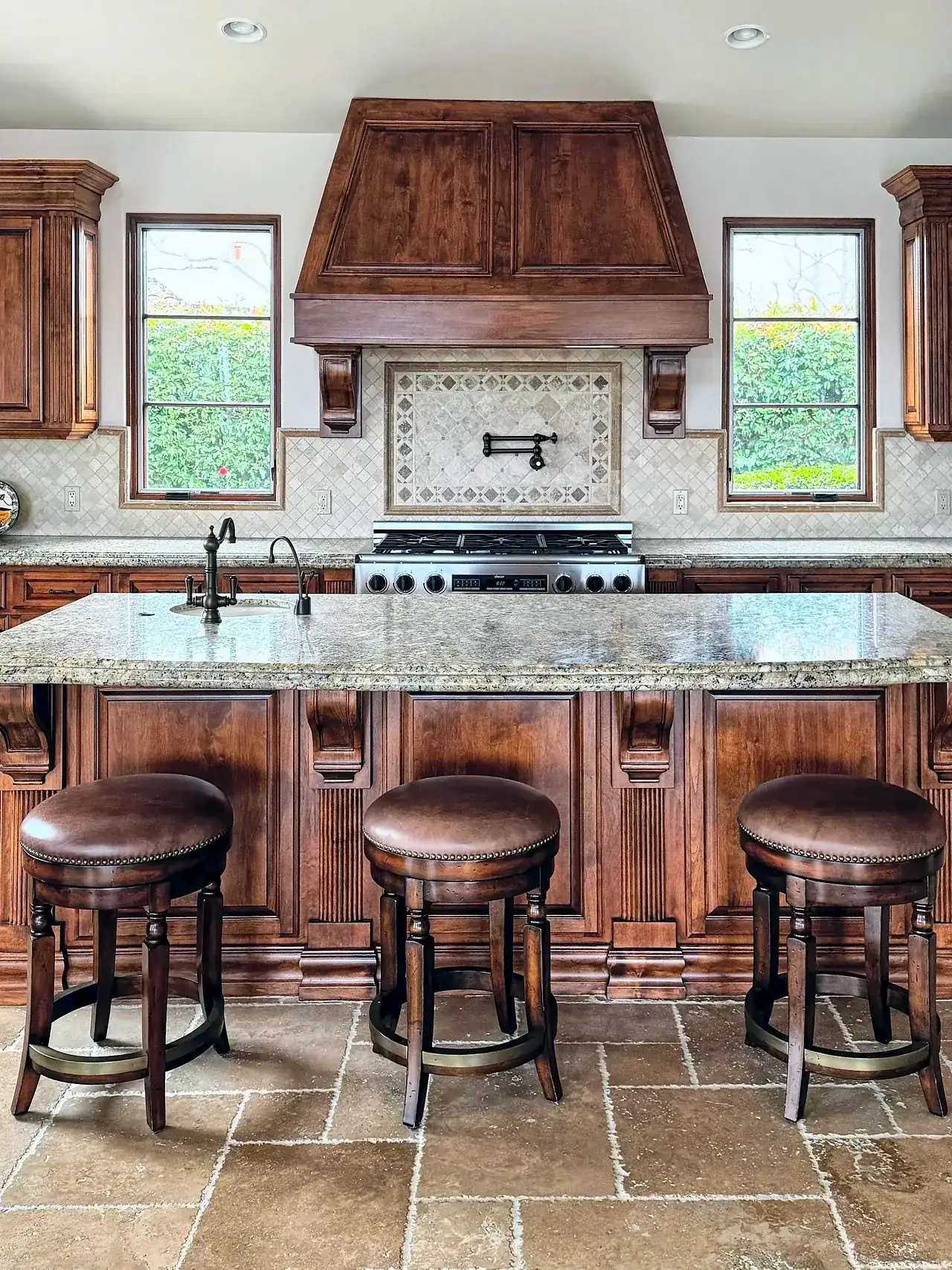 Santaluz kitchen before the remodel, with dark stained cabinets, a wood range hood, tumbled stone floors and a granite island with three counter stools