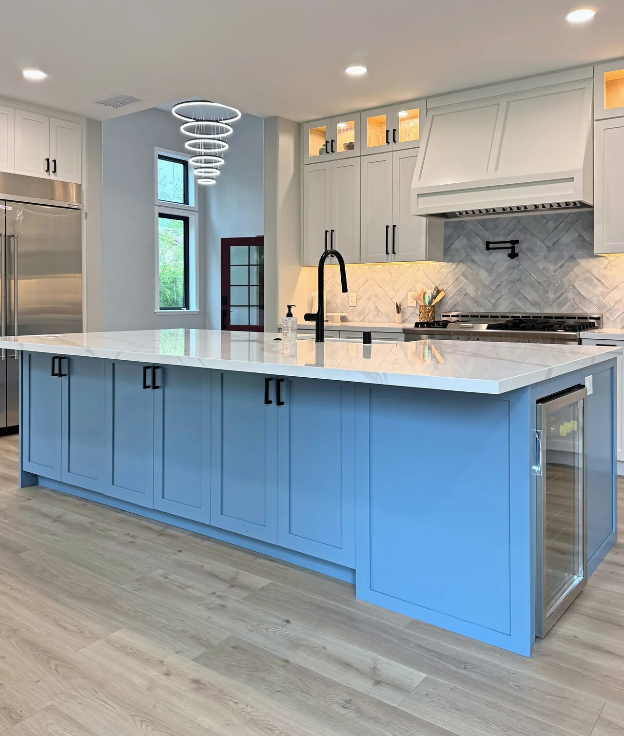 Kitchen with a long blue shaker island and a white quartz top, the range wall with a herringbone backsplash and painted hood, and lit glass-front uppers