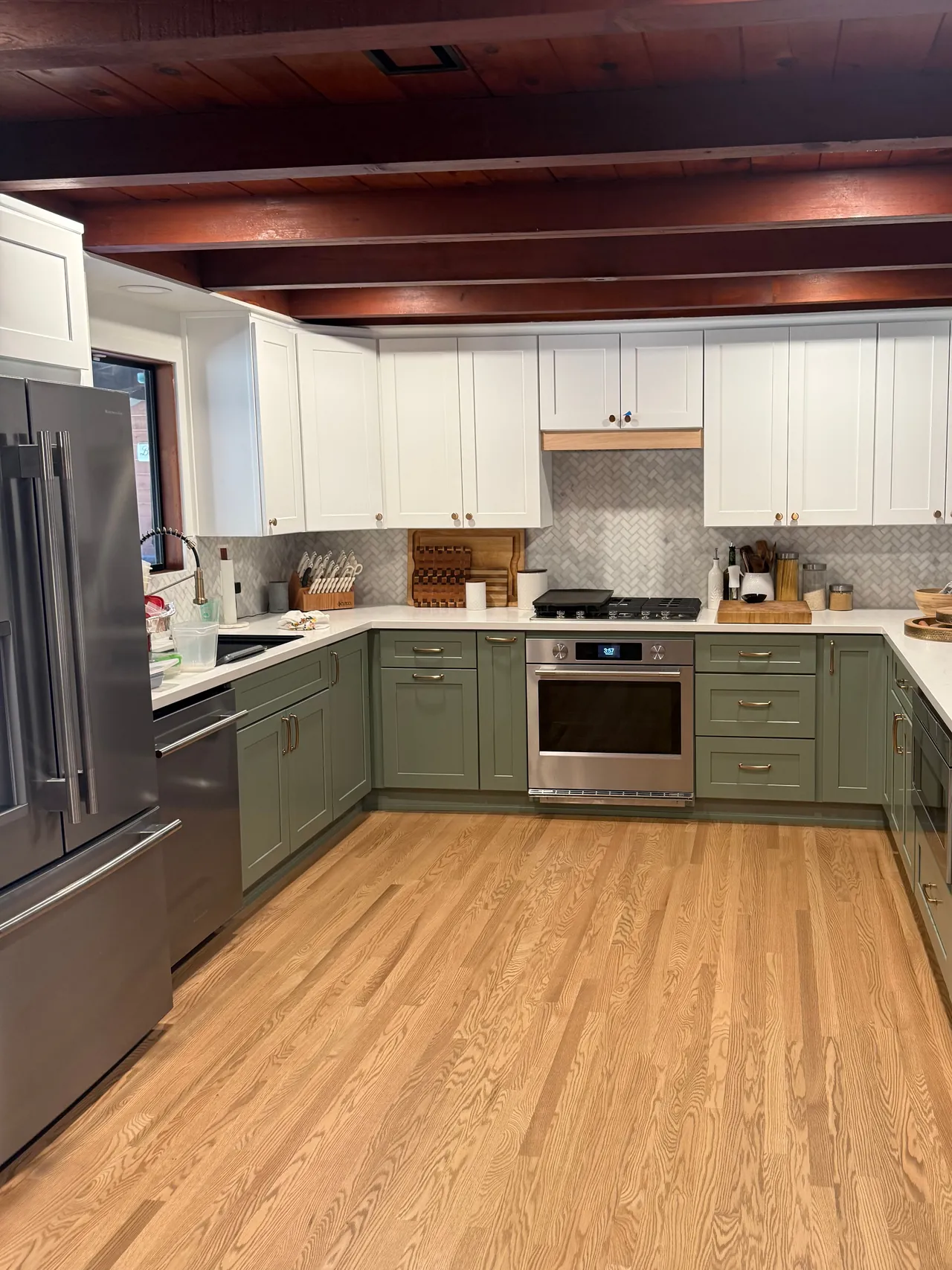 Kitchen with sage green lower cabinets, white shaker uppers, a marble herringbone backsplash and oak floors under a wood beam ceiling