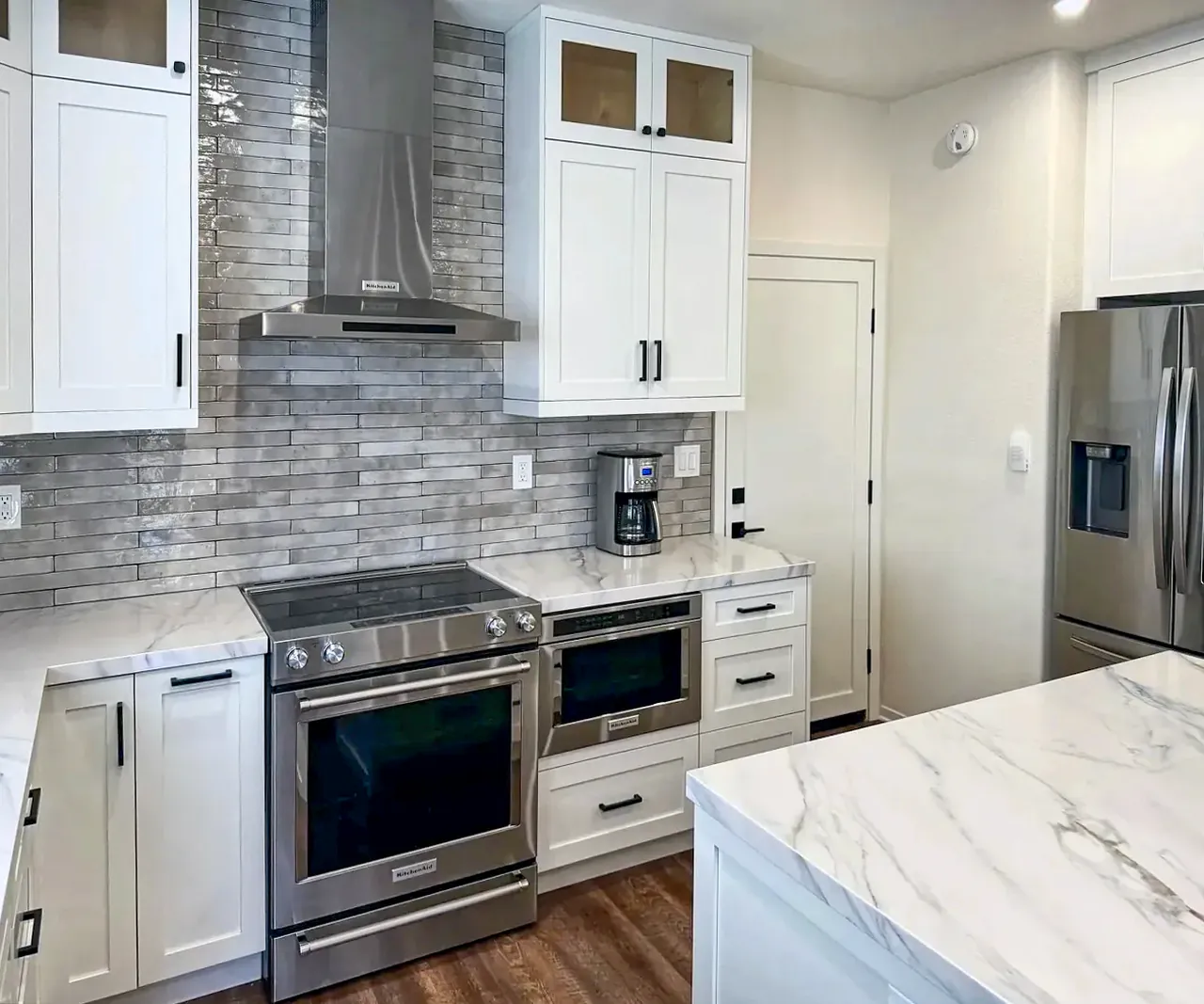 The finished Oceanside kitchen, with white shaker cabinets, a slide-in range, a chimney hood and a gray glossy tile backsplash