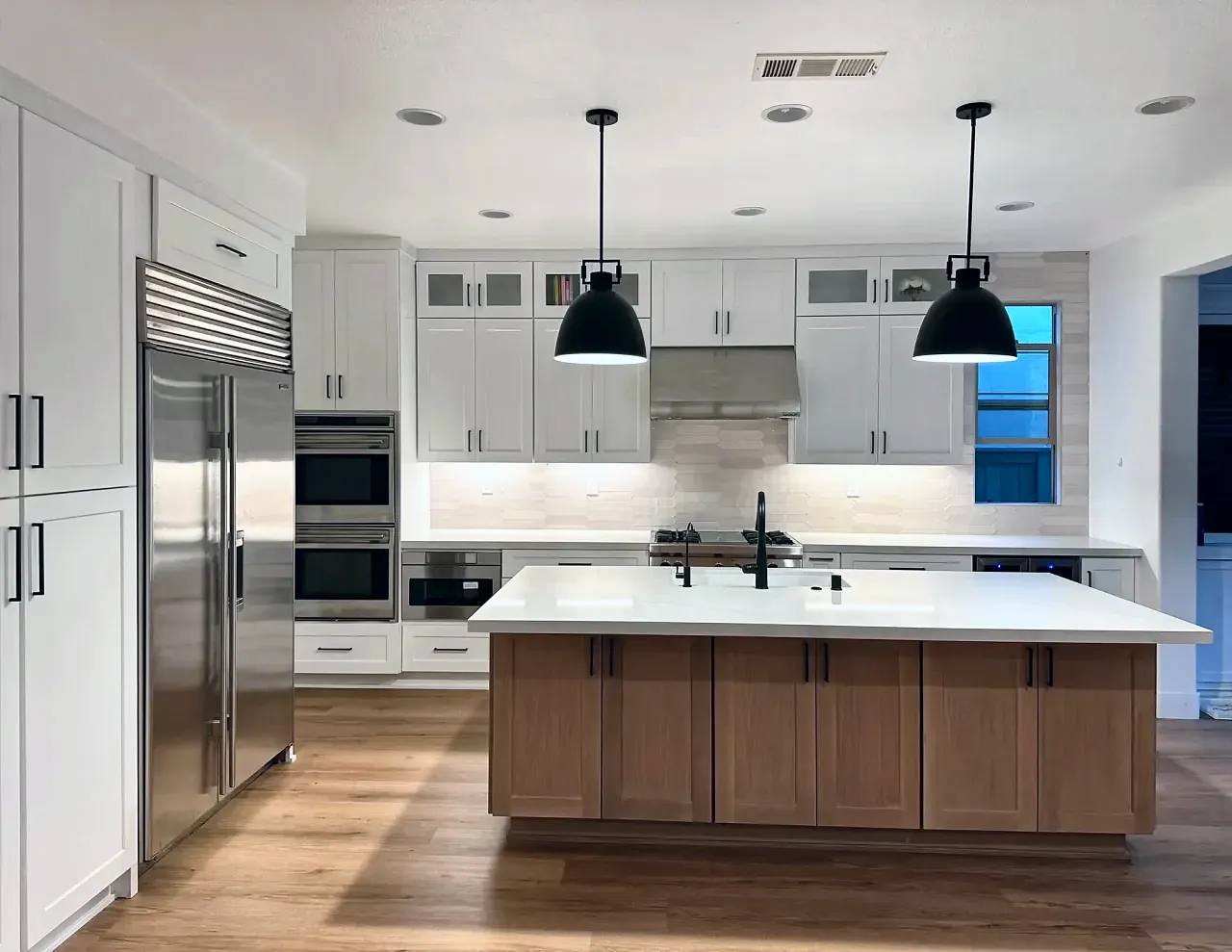 The same white shaker kitchen: the stained wood island under two black pendants, the range wall, and glass-door cabinets above the uppers