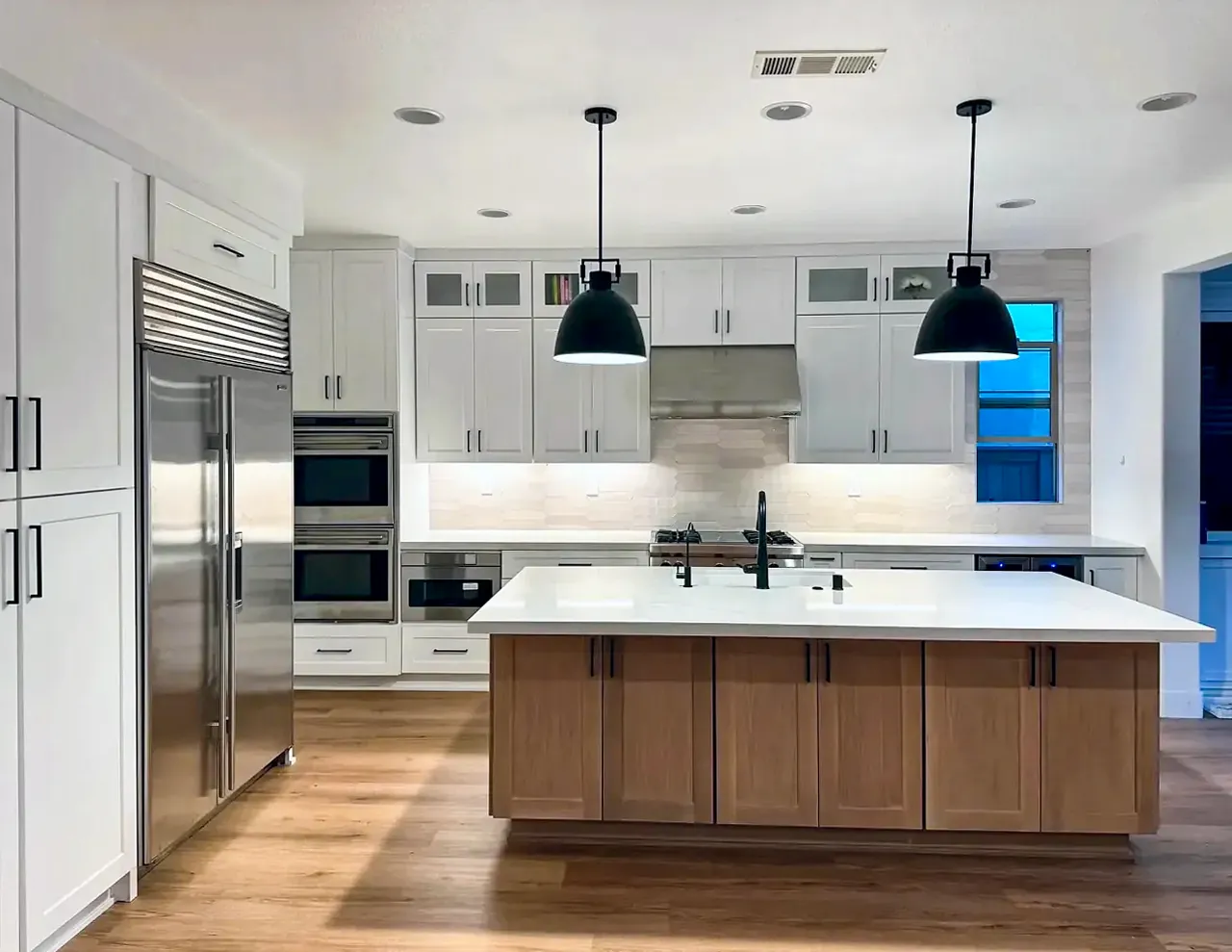 The same white shaker kitchen: the stained wood island under two black pendants, the range wall, and glass-door cabinets above the uppers