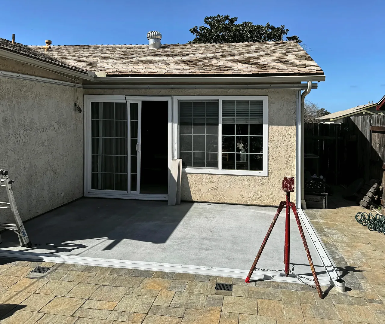 A new concrete slab poured in the corner between two walls of the house, ready for the sunroom walls, with a laser level set up on the edge