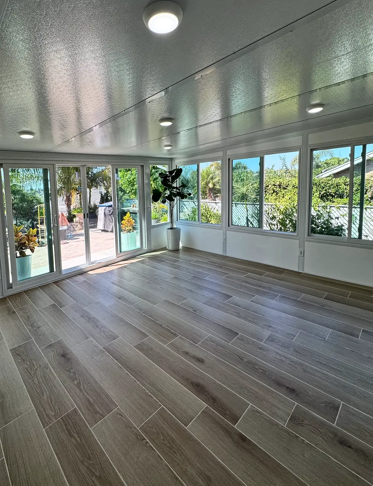Inside the finished sunroom, with round ceiling lights, a four-panel sliding glass door and windows on two sides over a low white wall, and a wood-look tile floor