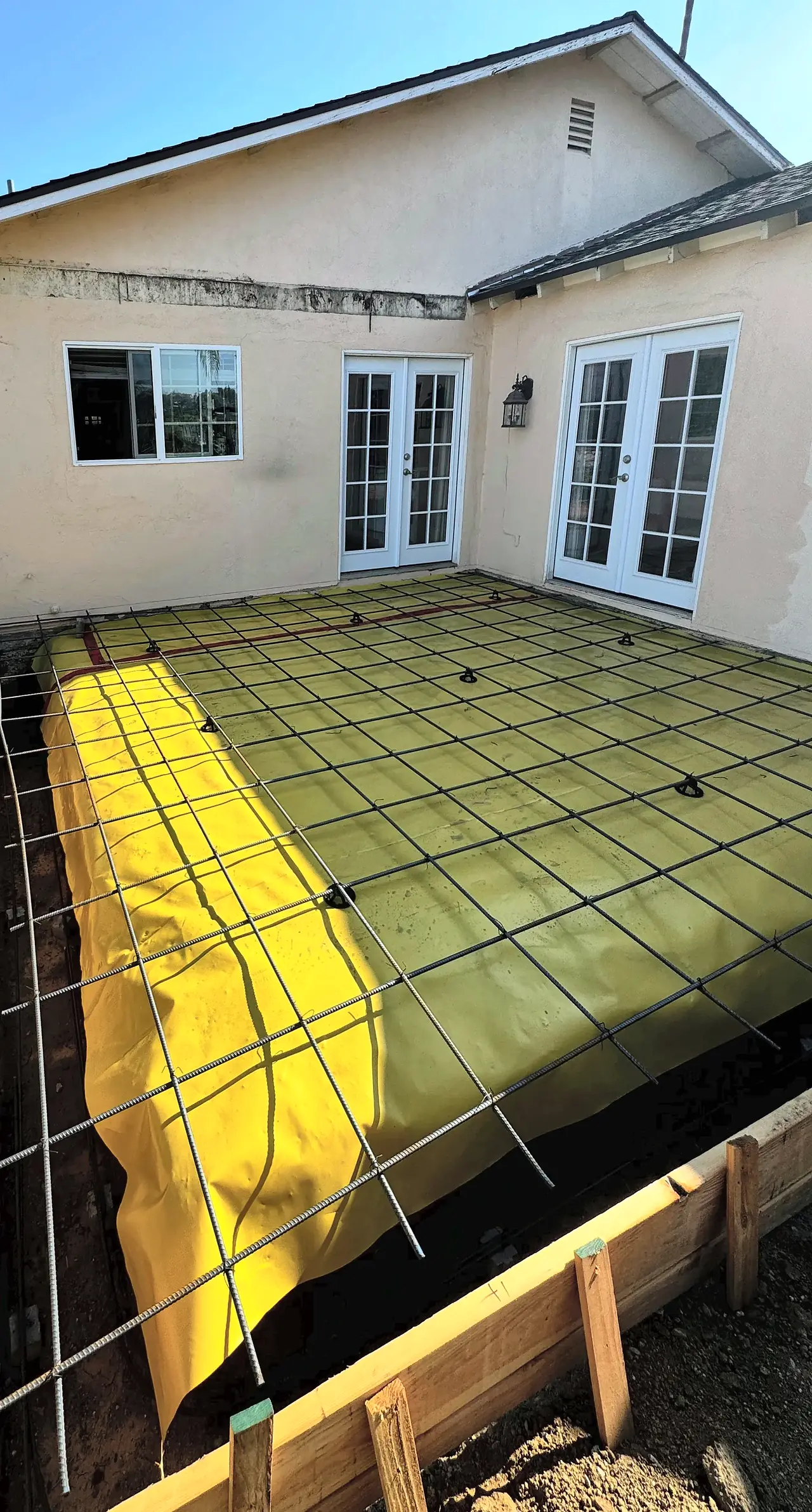 Wood forms, a yellow vapor barrier and a rebar grid set in the corner behind the house, ready for the new slab